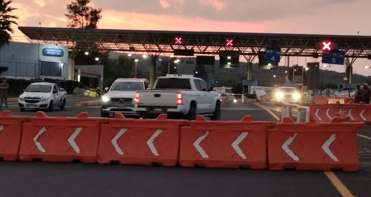 Valla de contención naranja colocada para bloqueas las casetas de cobro en Michoacán con camionetas y automóviles blancos detrás durante la tarde