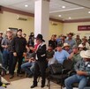 Una reunión de productores o agricultores de maíz (muchos con sombreros vaqueros) en un salón interior. Un hombre con sombrero negro y traje oscuro