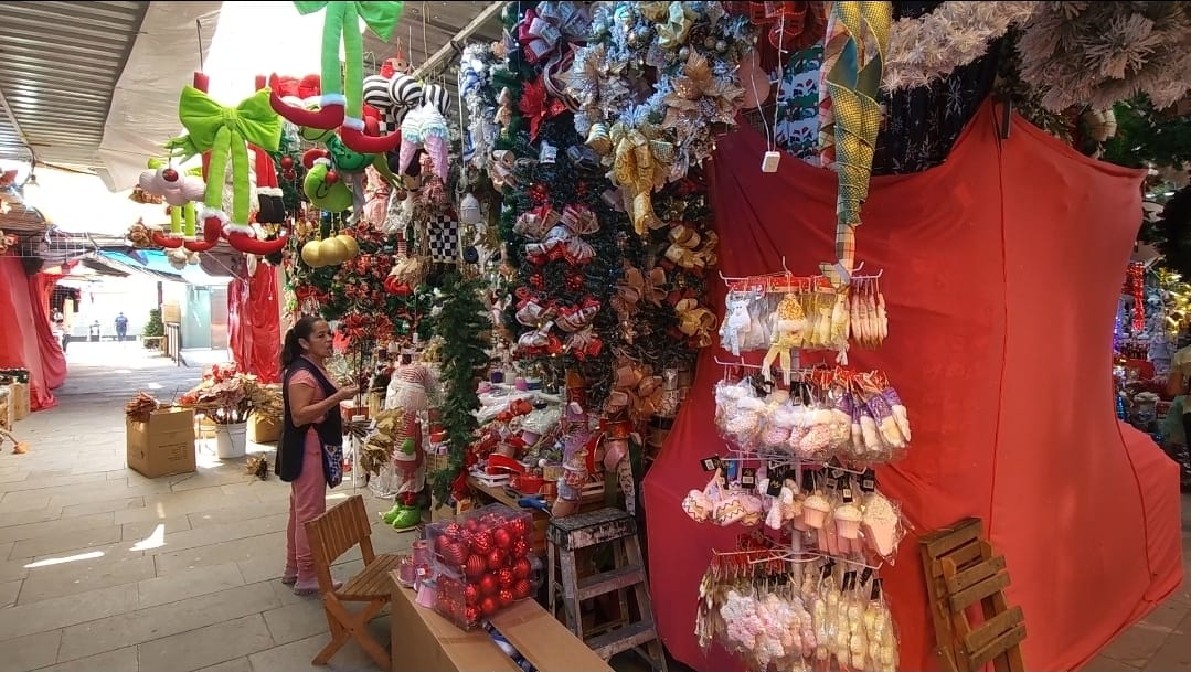 Una mujer de espaldas mira un saturado puesto de mercado lleno de adornos navideños de color verde, rojo y blanco, con luces y muñecos colgantes en el pasillo d