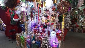 Un puesto de mercado repleto de adornos navideños de colores brillantes y luces. Se ven guirnaldas, coronas, esferas de colores apiladas en tubos de plástico