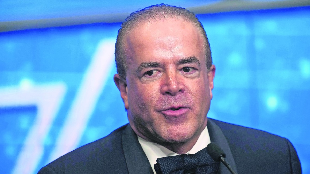 Primer plano de un hombre calvo, Raúl Cantú de la Garza (político), con traje y pajarita negra, hablando seriamente en un evento contra un fondo azul brillante