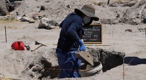 Hombre con sombrero y cubrebocas cavando y echando tierra en una cubeta con una pala dentro del Rancho Izaguirre en Teuchitlán