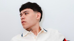 Primer plano de Junior H (cantante de corridos tumbados) con un corte de cabello estilo mullet corto, mirando hacia un lado. Lleva una camisa blanca con cuello