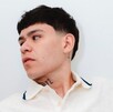 Primer plano de Junior H (cantante de corridos tumbados) con un corte de cabello estilo mullet corto, mirando hacia un lado. Lleva una camisa blanca con cuello