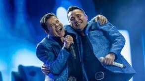 El vocalista de La Arrolladora Banda El Limón, Jorge Medina, abraza a un compañero en el escenario. Ambos llevan chaquetas azules brillantes y cantan sonrientes