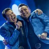 El vocalista de La Arrolladora Banda El Limón, Jorge Medina, abraza a un compañero en el escenario. Ambos llevan chaquetas azules brillantes y cantan sonrientes