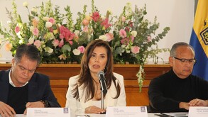 Una mujer (Verónica Delgadillo) hablando en un micrófono, flanqueada por dos hombres (Ramiro López y Héctor Raúl Gómez), sentados ante un gran arreglo floral