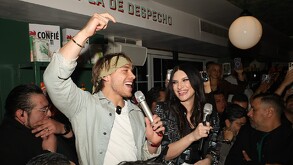 El influencer Aldo De Nigris (hijo, de chaqueta verde menta y pañuelo) sostiene un micrófono y grita junto a una mujer con chaqueta de lentejuelas.