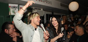 El influencer Aldo De Nigris (hijo, de chaqueta verde menta y pañuelo) sostiene un micrófono y grita junto a una mujer con chaqueta de lentejuelas.