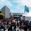 Una gran multitud de personas (visitantes de la FIL) afuera del edificio de la Expo Guadalajara bajo el sol. El edificio principal tiene una carpa blanca grande