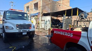 Una grúa blanca y una camioneta roja de BOMBEROS están en la escena de un incendio. El fuego ha quemado un vehículo dentro de un garaje improvisado anexo