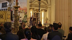 Una vista trasera de una congregación sentada en una iglesia de estilo barroco. Un cardenal (de sotana morada) está de pie en el púlpito dando misa.