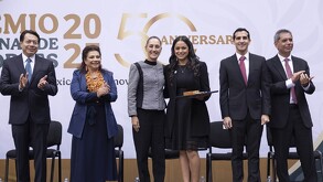 Una fila de seis personas vestidas de forma formal en el escenario. Una mujer en el centro recibe un premio de otra mujer (posiblemente Claudia Sheinbaum), cele