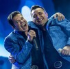 El vocalista de La Arrolladora Banda El Limón, Jorge Medina, abraza a un compañero en el escenario. Ambos llevan chaquetas azules brillantes y cantan sonrientes