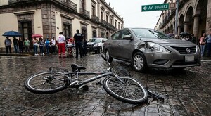Bicicleta tumbada sobre un suelo adoquinado al lado de un automóvil Nissan gris con impacto en la parte delantera. Paramédicos, policías y personas alrededor