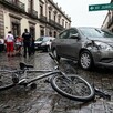 Bicicleta tumbada sobre un suelo adoquinado al lado de un automóvil Nissan gris con impacto en la parte delantera. Paramédicos, policías y personas alrededor