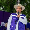 El político Carlos Manzo (con sombrero de paja y chaleco púrpura) habla en un micrófono en un evento al aire libre en un parque. Otros asistentes con sombreros