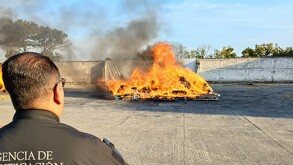 Un oficial de la Agencia de Investigación de espaldas observa una gran hoguera. Grandes llamas y humo oscuro se levantan de la quema de droga incautada
