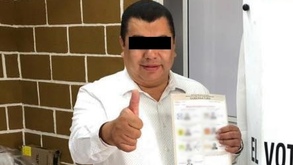Un hombre vestido de camisa blana sosteniendo una boleta electoral y haciendo una seña con la mano.