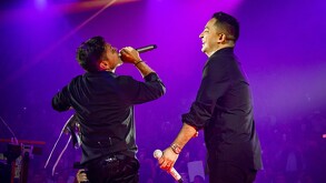 El cantante Jorge Medina (espalda) y un compañero cantan juntos en un escenario de concierto. Ambos visten camisas negras y están bajo una intensa luz púrpura,