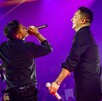 El cantante Jorge Medina (espalda) y un compañero cantan juntos en un escenario de concierto. Ambos visten camisas negras y están bajo una intensa luz púrpura,