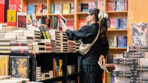 Una mujer de espaldas con ropa negra y anteojos, con un moño blanco grande en el cabello, navega por pilas de libros en los estantes de un expositor