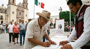 Señor con sombrero firmando documentos de registro para la Pensión Bienestar con un hombre atendiéndolo y otros adultos mayores haciendo fila detrás