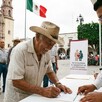 Señor con sombrero firmando documentos de registro para la Pensión Bienestar con un hombre atendiéndolo y otros adultos mayores haciendo fila detrás