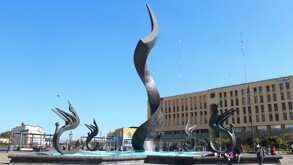 Una moderna escultura metálica en forma de flama en una fuente circular, rodeada de esculturas de aves. En el fondo, se ve un edificio de color crema bajo un ci