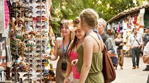 Tres mujeres (turistas) sonrientes examinan un expositor lleno de gafas de sol, sombreros y souvenirs artesanales en un mercado al aire libre de Puerto Vallarta