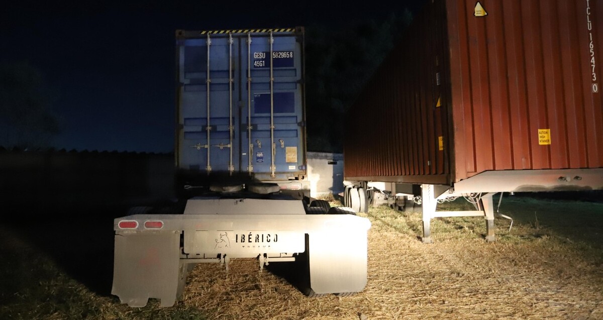Parte trasera de un tractocamión color azul junto a un tráiler color rojo, ambos estacionados en un terreno con pasto y tierra