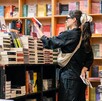 Una mujer de espaldas con ropa negra y anteojos, con un moño blanco grande en el cabello, navega por pilas de libros en los estantes de un expositor
