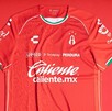 Primer plano de la nueva camiseta de fútbol de local del Atlas (roja). Tiene patrocinadores como Caliente.mx y Charly, y detalles en blanco y verde oscuro