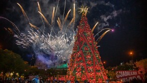 Gran árbol de Navidad cónico y alto, decorado con luces, listones rojos y moños, iluminado por la noche en la plaza. En el cielo oscuro estallan pirotecnia