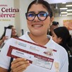 Alumna de secundaria con lentes viendo de frente y sonriendo mientras sostiene un sobre del Banco del Bienestar con personas de fondo
