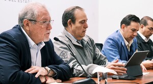 Autoridades de Jalisco sentados detrás de una mesa con micrófonos y una tablet delante de medios de comunicación con un fondo blanco