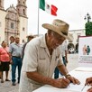 Señor con sombrero firmando documentos de registro para la Pensión Bienestar con un hombre atendiéndolo y otros adultos mayores haciendo fila detrás