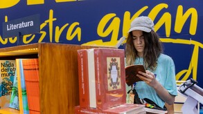 Una joven con gorra de béisbol y cabello largo lee un libro mientras se apoya en un estante de madera en la FIL (Feria Internacional del Libro). Un cartel super