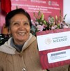 Una mujer sonriente, probablemente una adulta mayor, sostiene un sobre que contiene la Tarjeta del Banco del Bienestar con el logo del Gobierno de México.