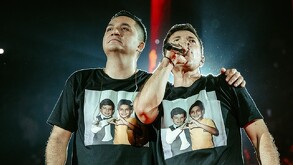 Se observa a dos hombres con playera negra, uno está cantando con micrófono en mano.