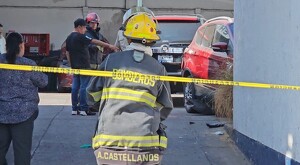 Bombero de Guadalajara de espaladas y detrás de una cinta policiaca ingresando a un taller mecánico donde hay automóviles y otras personas de pie