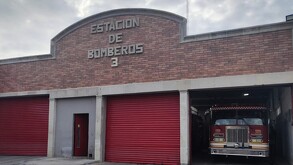 Se observa una fachada que dice Estación de Bomberos y se ve un camión de bomberos al lado.