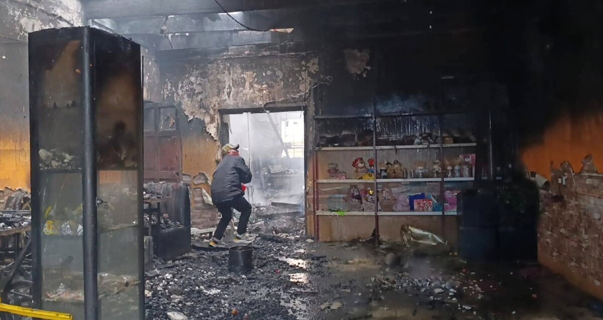 Interior de un restaurante con estanterías y diversos objetos calcinados. Una persona arrojando agua a las paredes quemadas