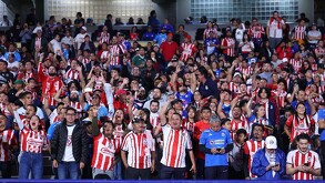 Una gran multitud de aficionados de Chivas (la mayoría vistiendo camisetas de rayas rojas y blancas) animan ruidosamente desde las gradas de un estadio