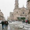 Calle y edificios de Lagos de Moreno con agua nieve cayendo y el cielo nublado. Personas caminando abrigadas, algunas con sombrillas y la catedral de fondo