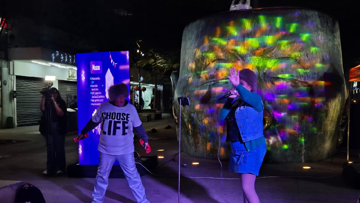 Una mujer canta en un micrófono frente a la escultura de una cabeza gigante iluminada con luces de colores proyectadas.