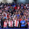 Una gran multitud de aficionados de Chivas (la mayoría vistiendo camisetas de rayas rojas y blancas) animan ruidosamente desde las gradas de un estadio