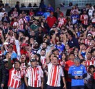 Una gran multitud de aficionados de Chivas (la mayoría vistiendo camisetas de rayas rojas y blancas) animan ruidosamente desde las gradas de un estadio