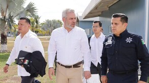 El político Alfredo Ramírez Bedolla (centro, camisa blanca) camina junto a un hombre en un uniforme policial y otros asistentes bajo un sol brillante