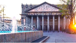 El histórico Teatro Degollado en Guadalajara. La fachada neoclásica tiene columnas y un relieve. Una fuente moderna con chorros iluminados en azul